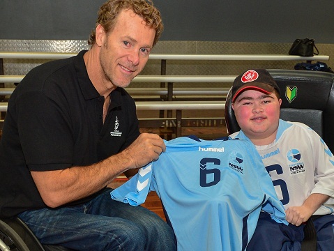 Captain of the NSW Team is Chris Suffield, here receiving his jersey from John MacleanThe New South Wales Powerchair Football received their Team jerseys today at a special training session today.Starting on Tuesday 5th August the NSW Team will represent the State at the Powerchair National Titles, held at Kevin Betts Stadium , Mount Druitt. Competing over the tournament are teams from other states along with a representative team from New Zealand. The Tournament concludes on Saturday 9th August, with a Grand Final. Followed by the Ditch Cup, where the newly named Australian Team will compete against the New Zealand Team.In attendance to present each player with their jersey was John Maclean. John is an Australian triathlete, rower, and motivational speaker. A promising rugby league player in his youth, he became a paraplegic after being knocked from his bicycle by a truck in 1988. He became the first person in a wheelchair to complete the Ironman World Championship and swim the English Channel in 1995 and 1998, respectively.This year’s Captain of the team is Chris Suffield of Central Coast Mariners. At the conclusion of the presentation the team lined up for a team photo, including Team Staff. Rear Row: (Left to Right) Andrew Scollard (WSW, Team Mechanic); Peter Turnbull (Mariners, Coach); Robbie Wright (Volunteer, Asst Coach) Front Row: (L to R) John Maclean; Alex Scollard (WSW); Jacob Cross (WSW); Ben Keyte (WSW); James Kim (Newcastle Jets); Jordan Crane (Sydney FC); Chris Turnbull (Mariners) &amp; Chris Suffield Capt (Mariners).